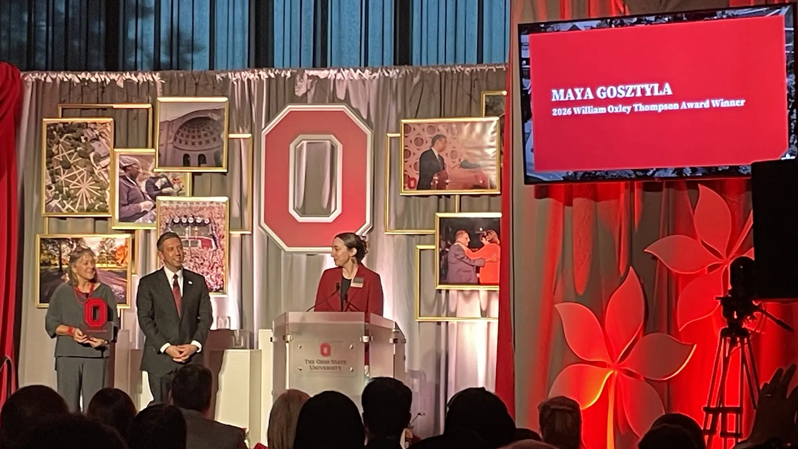 Maya standing at a podium to speak at the awards ceremony. Everything is scarlet and grey and full of Block O's